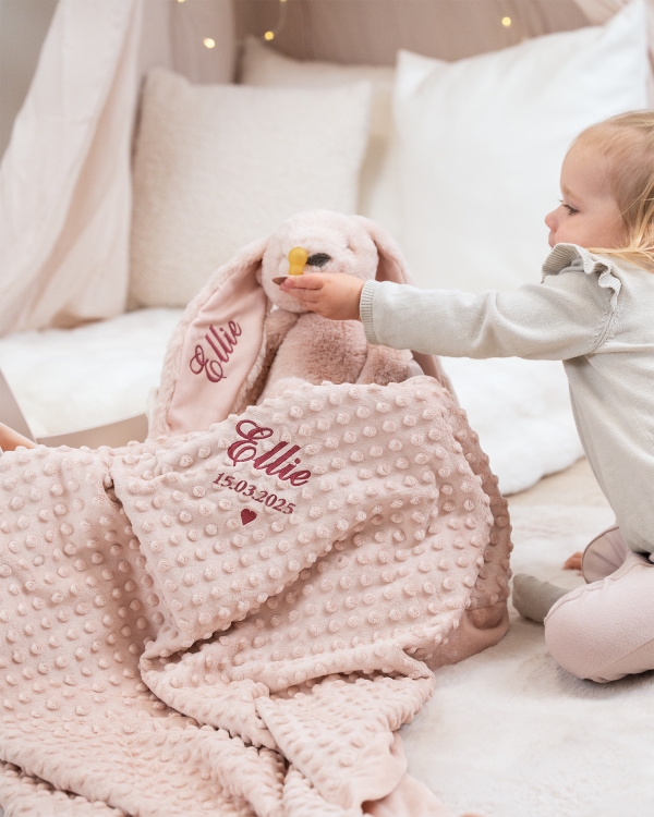 Vaaleanpunainen peitto ja pehmolelu-kani, johon on kirjailtu nimi ja syntymäaika