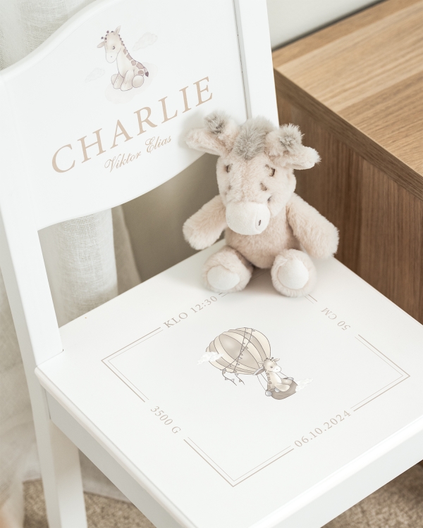 Valkoinen kastetuoli, jossa nimi selkänojassa sekä päivämäärä, aika ja kirahviaihe istuimessa