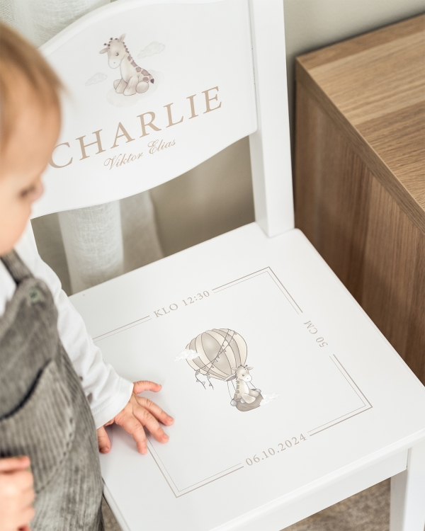 Valkoinen kastetuoli, jossa nimi selkänojassa sekä päivämäärä, aika ja kirahviaihe istuimessa