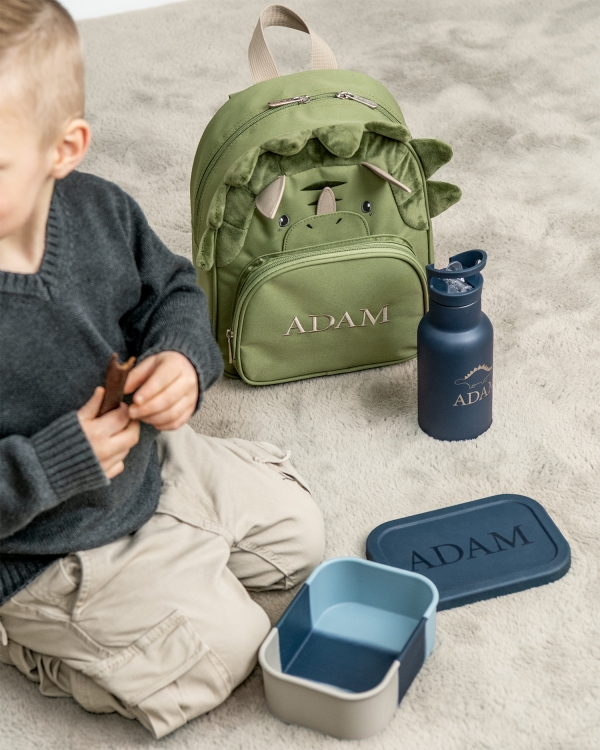 Lasten dinosauruksen muotoinen reppu, jossa on kirjailtu nimi ja sininen lounasrasia ja vesipullo